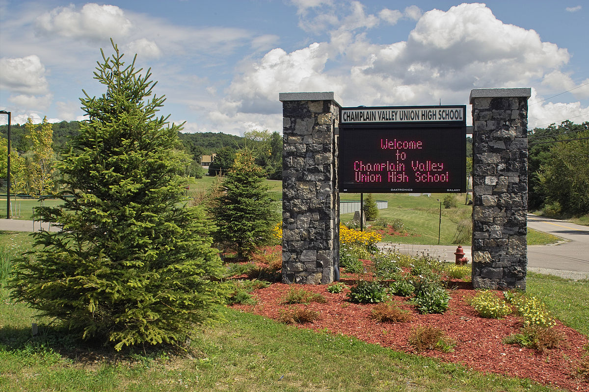 Champlain Valley Union High School sign in a landscaped area with a stone base, surrounded by trees and plants, displaying a welcome message.