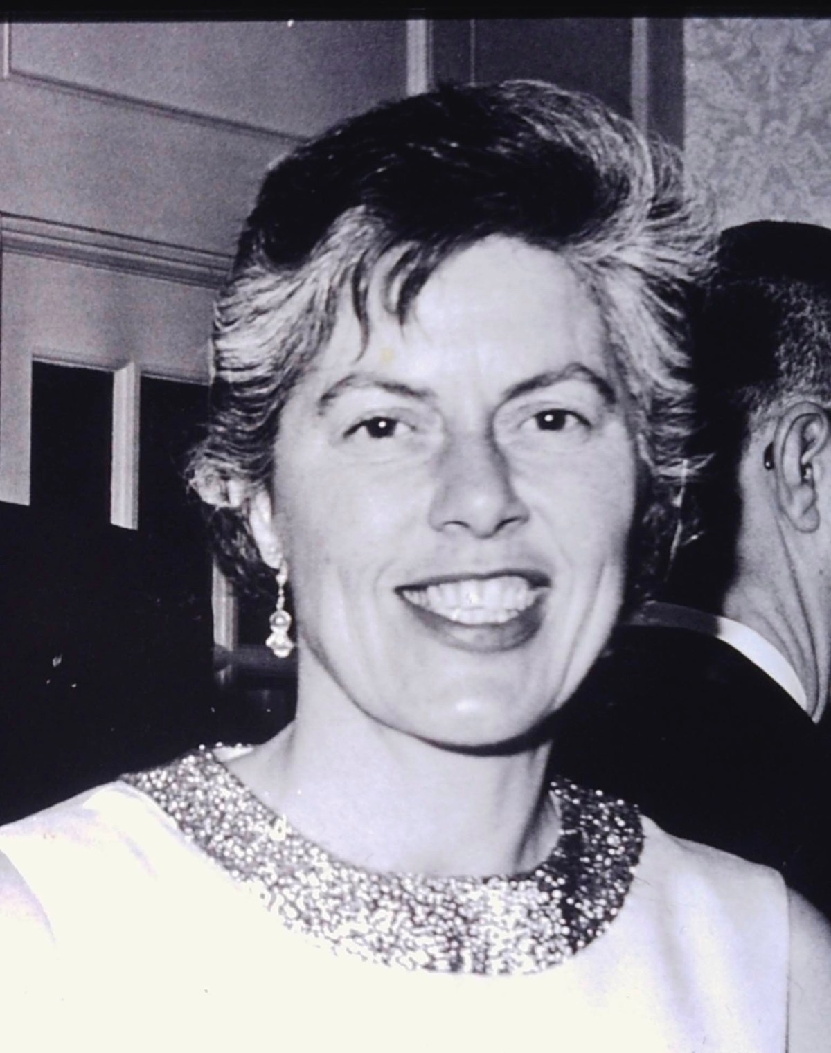 Black and white photo of a woman smiling, wearing a dress with a sequined collar and drop earrings, standing indoors beside another person.