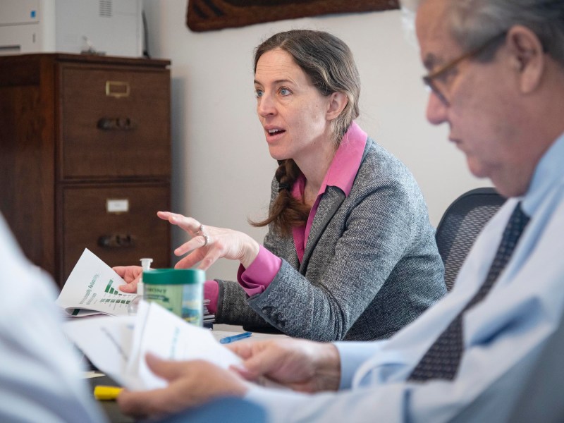 A woman in a gray blazer talks during a meeting, gesturing with her hand. Two men, one blurred in the foreground and the other beside her, are seated, holding papers. A filing cabinet is in the background.