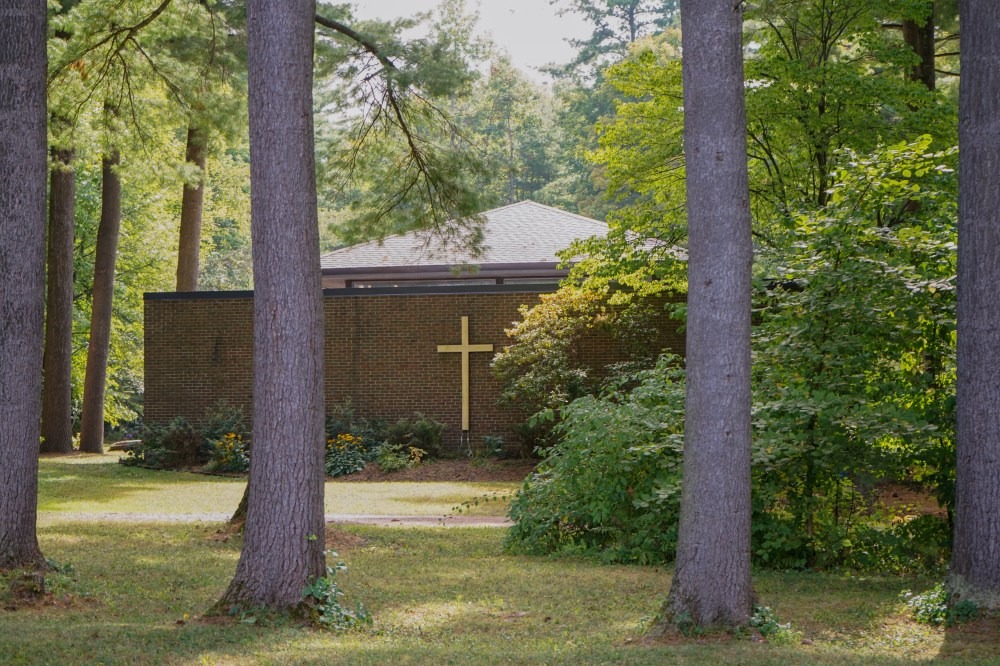 A brick church with a large gold cross on its facade is nestled among tall pine trees and surrounded by lush greenery.