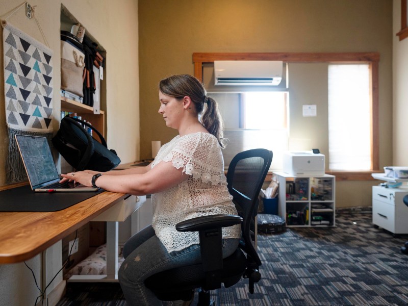 A woman sits at a desk working on a laptop in a home office with shelves, a printer, and office supplies visible in the background.