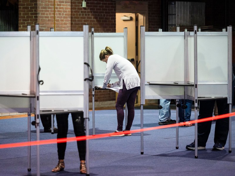 People standing at voting booths in a voting station with a red tape barrier in the foreground.