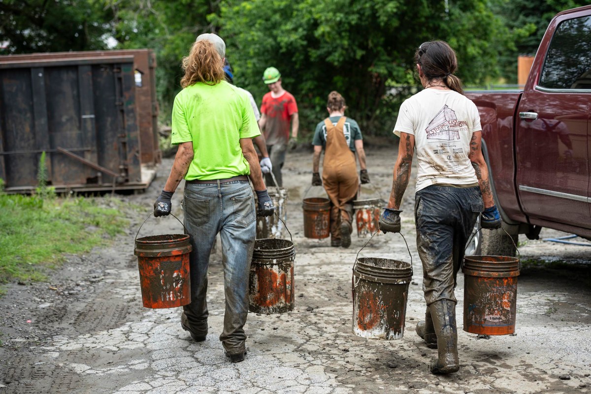 Several people are walking on a muddy path, carrying dirty buckets. They are wearing stained work clothes and boots. Trees and a truck are visible in the background.