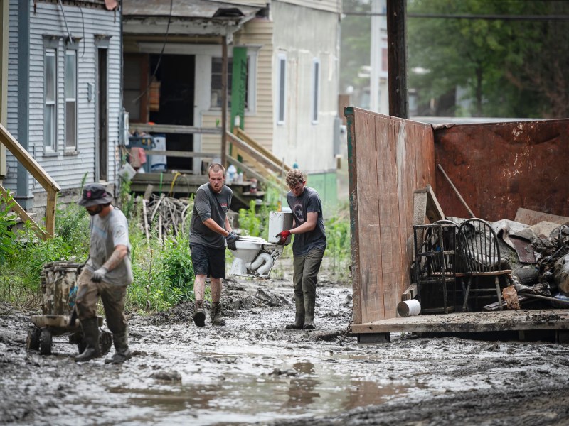 Three people working in a muddy, flooded alleyway clean up and remove debris into a large dumpster. One person pushes a wheelbarrow, while the other two carry items through the sludge.