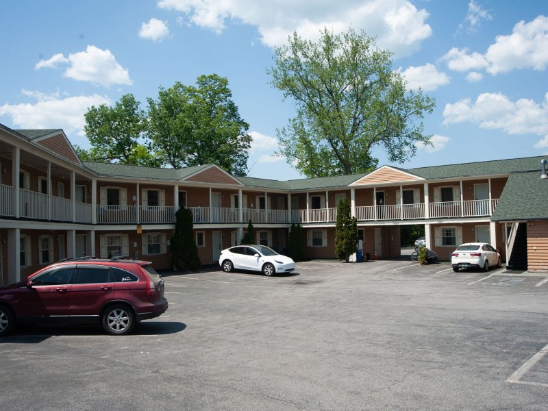 A parking lot with three cars in front of a two-story motel. The building has exterior staircases and balconies. Trees and clouds are in the background.