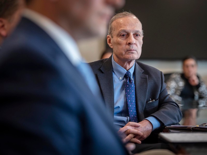 A man in a suit and tie sits attentively at a conference table with other individuals blurred in the foreground and background.