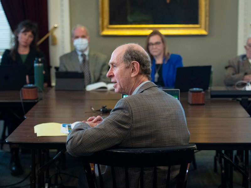 A man in a suit speaks at a table during a meeting with four other people, two using laptops. One person wears a mask.