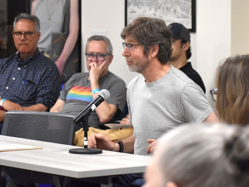 A man in a gray t-shirt speaks into a microphone at a meeting, while people sit and listen in the background.