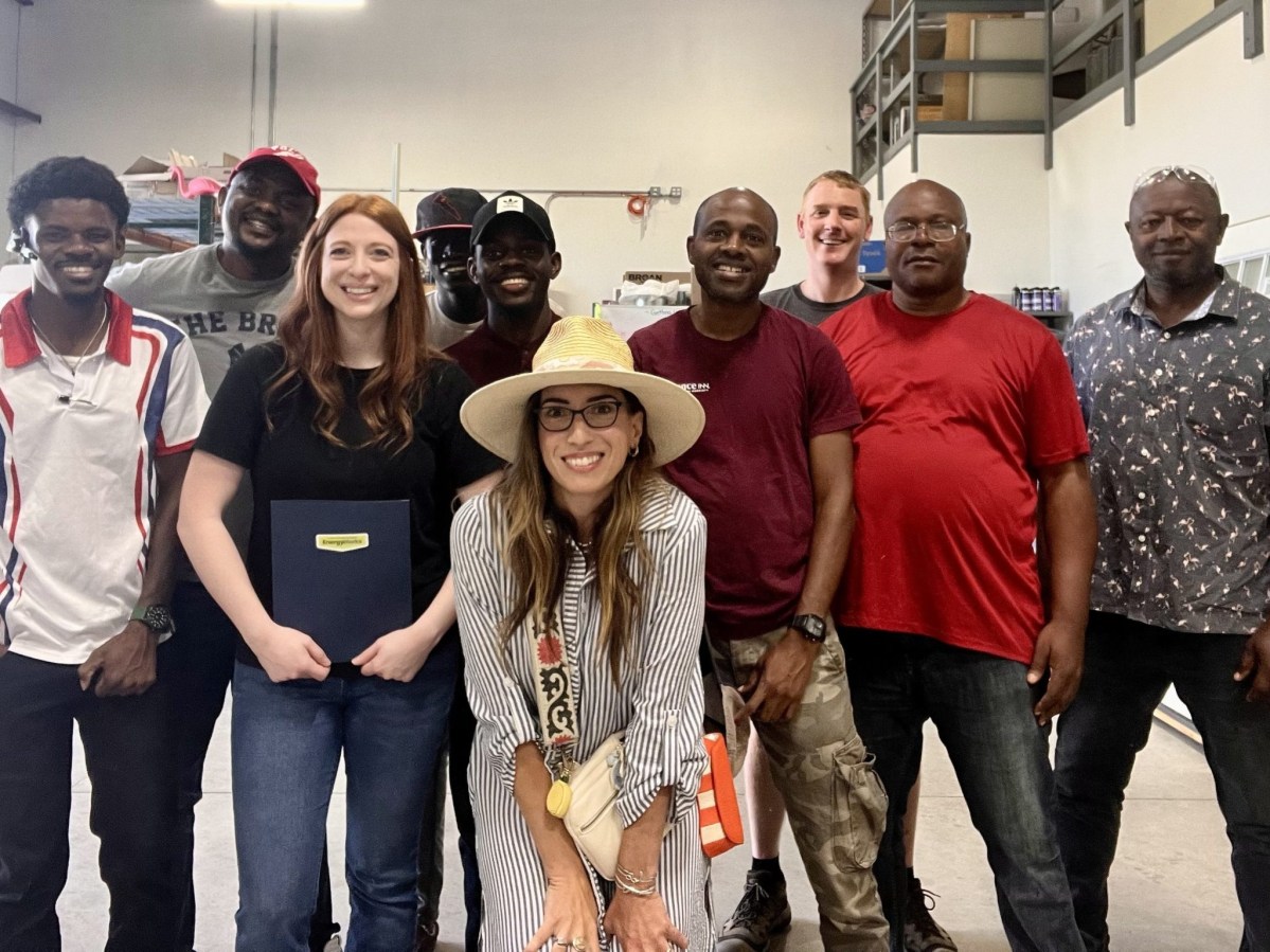 A group of nine adults, including one woman wearing a hat, poses and smiles for a photo inside an industrial or workshop setting.