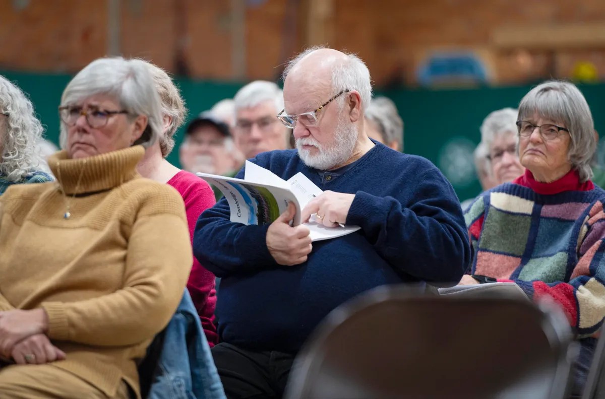 People seated indoors, looking attentive. A man in the middle reads a booklet. Casual, layered attire in autumn colors.