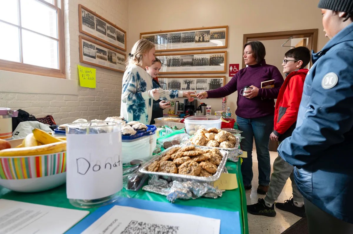 People at a bake sale table, exchanging money for food. Cookies, muffins, and bananas are displayed. A donation jar is also present.