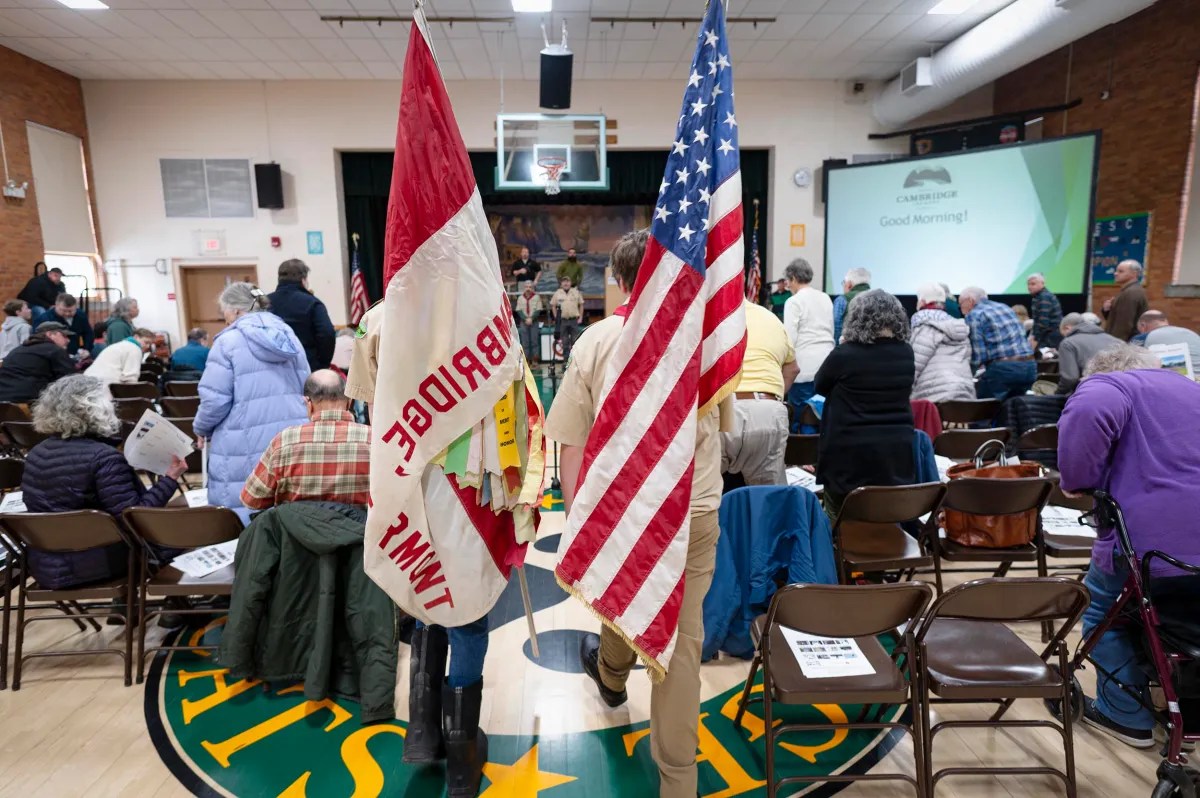 People attending an indoor community meeting, with visible flags of Bridgeport and the United States in the foreground. Seats are arranged in a school gymnasium.