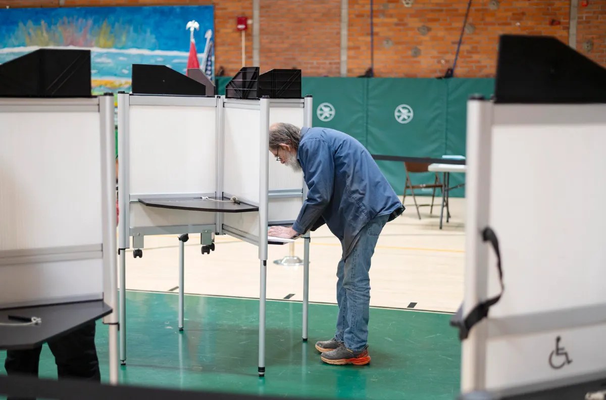 A person stands at a voting booth in a gymnasium, filling out a ballot. The setting includes a basketball court and green gymnasium mats.