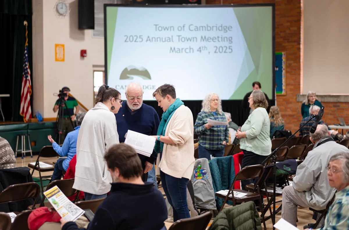 People gather and converse in a hall with a projection screen displaying "Town of Cambridge 2025 Annual Town Meeting March 4th, 2025" in the background.
