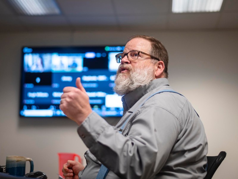 A man with glasses and a beard gestures with his thumb up while seated in front of a computer screen displaying various windows. He is wearing a gray shirt with suspenders.