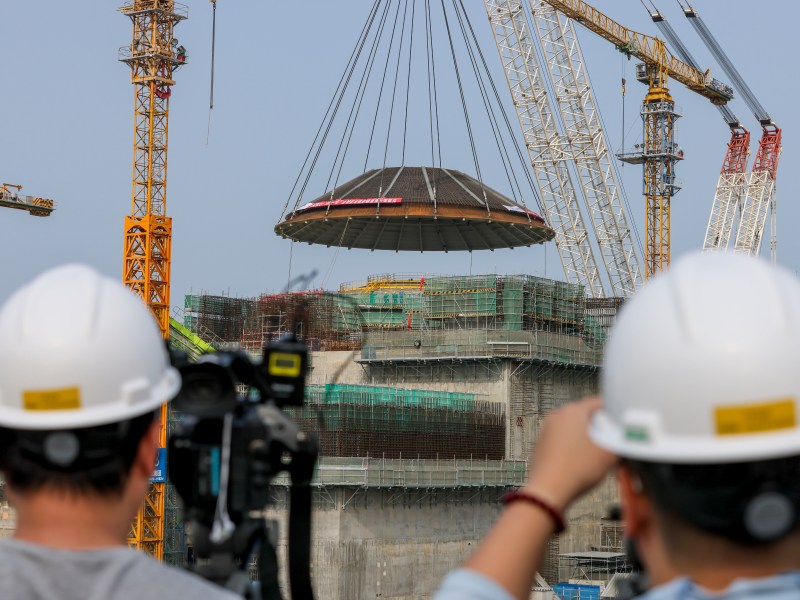 Workers monitor a large circular structure being lifted by cranes at a construction site, with two men in hard hats observing the process from the foreground.