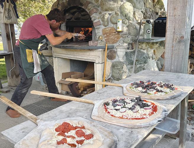 A person places a pizza into a stone pizza oven, with three uncooked pizzas topped with various ingredients on wooden peels on a table in the foreground.