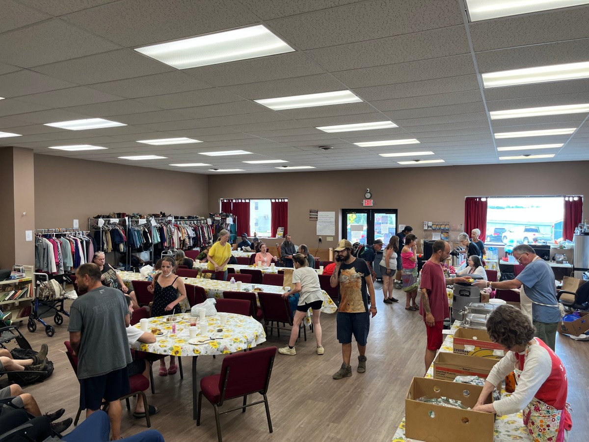 People gather in a community center, some sitting at tables and others organizing clothing and items. Clothing racks line the walls and boxes are open on tables.
