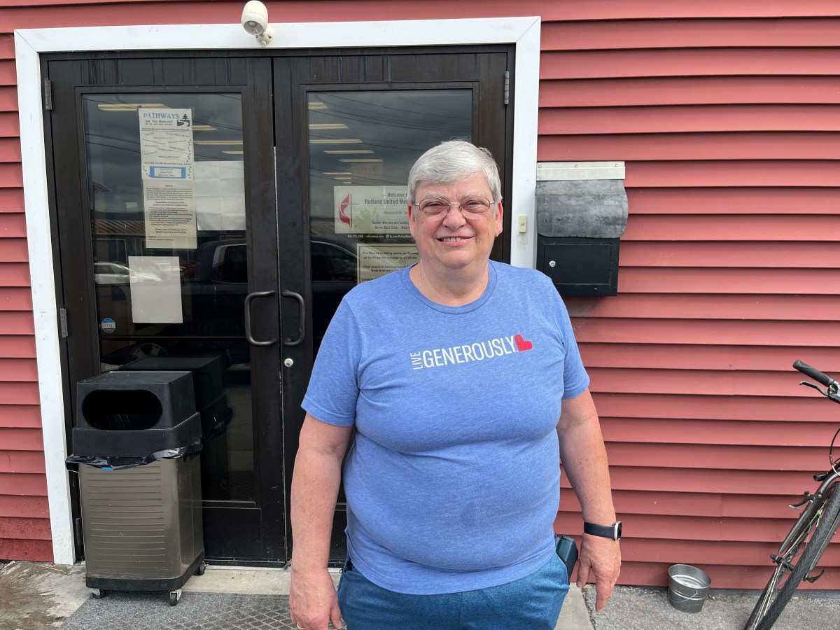 A person with short gray hair wearing a blue "Live Generously" T-shirt stands and smiles in front of a red building with glass doors.