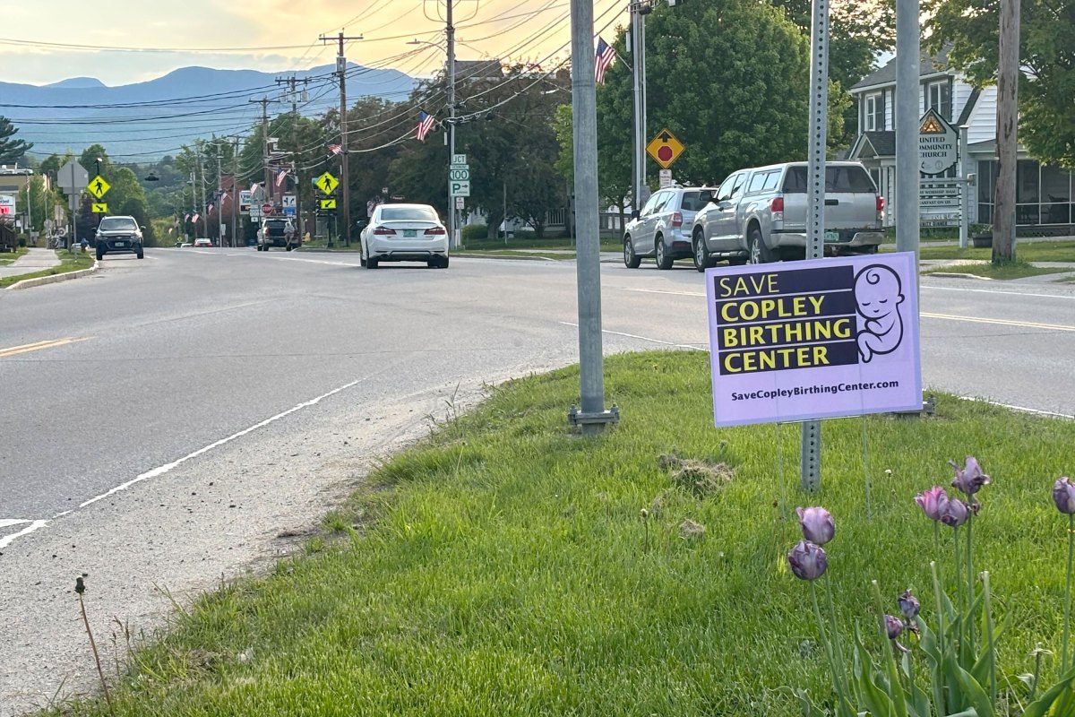 A "Save Copley Birthing Center" sign is displayed on a grassy roadside, with cars parked along the street and mountains visible in the background.
