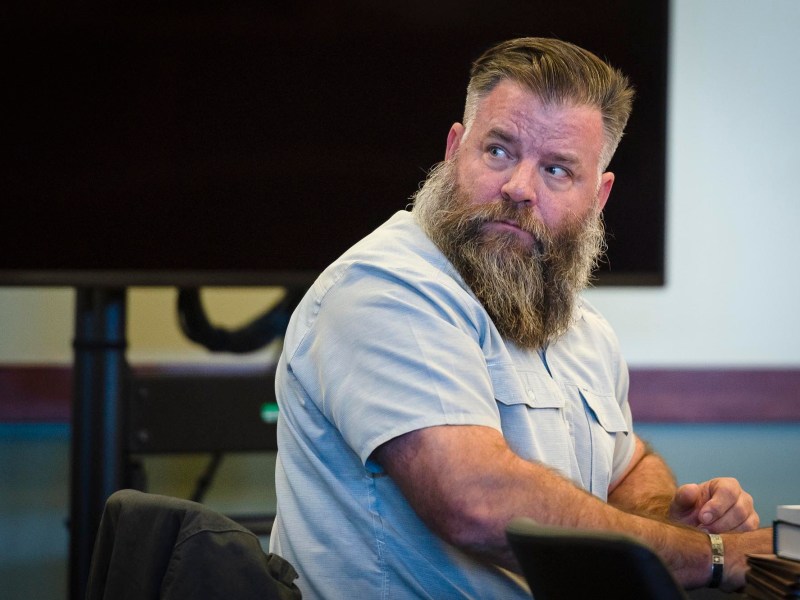 A man with a beard sitting in a courtroom.