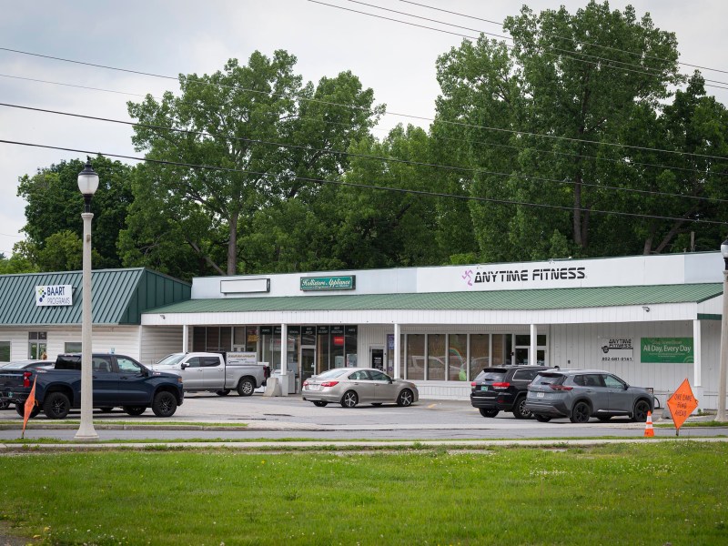 A small shopping plaza with parked cars features a fitness center, a market, and a laundromat, with trees in the background and an overcast sky.