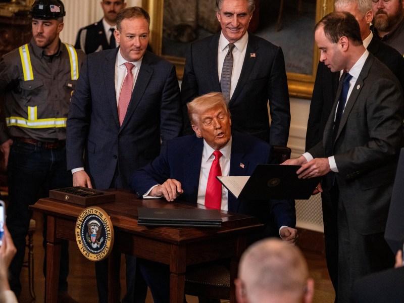 A man seated at a desk signs a document while others stand around him, including men in suits and workers in uniforms.