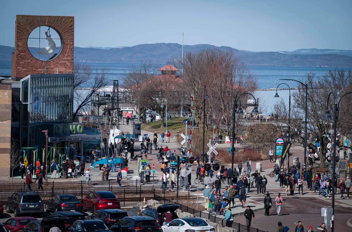 A bustling waterfront promenade with pedestrians, a large circular sculpture, and a view of a lake and hills in the distance.