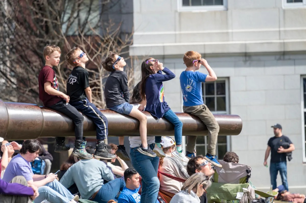Children sitting atop a metal structure, looking upwards with interest, while adults sit below.