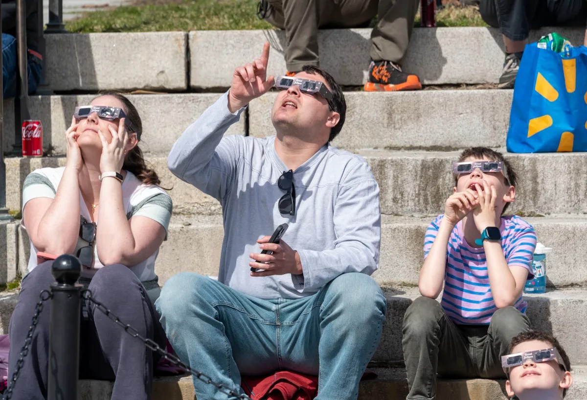 Group of people wearing protective glasses looking upwards in daylight, likely observing a solar eclipse or other celestial event.