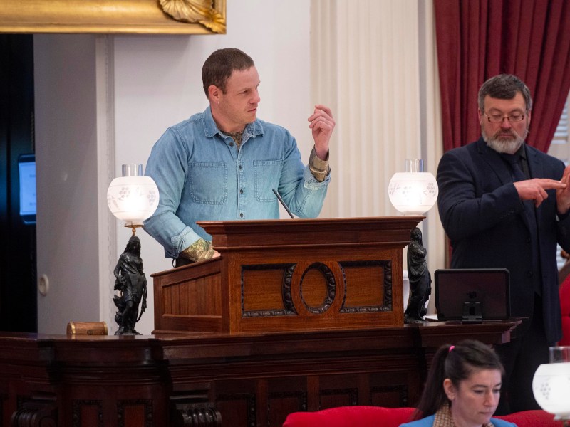 A man in a denim shirt speaks at a wooden podium in a formal room, while another man stands beside him using sign language.