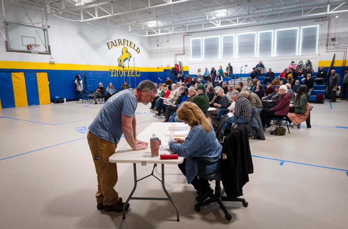 People gathered in a gymnasium for a community meeting. A man is standing at a table talking to woman seated. Attendees are seated on folding chairs.