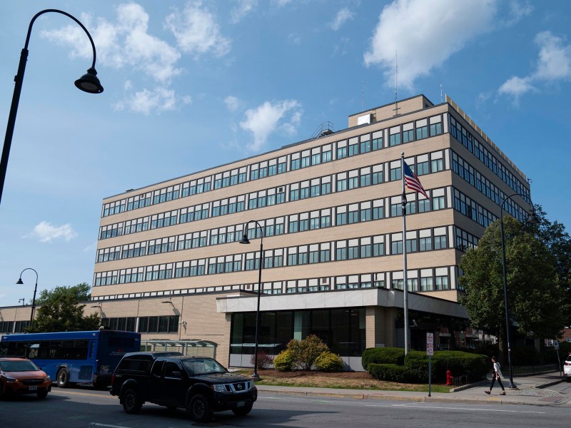 A large, rectangular office building with many windows, an American flag outside, cars and a blue bus parked nearby, and a partly cloudy sky overhead.