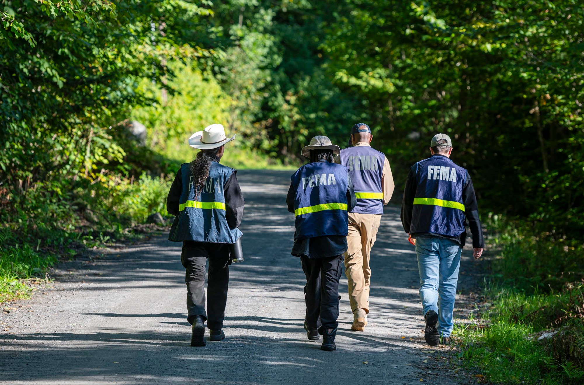 Four FEMA personnel walk down a gravel path surrounded by trees, wearing uniforms with "FEMA" on the back.