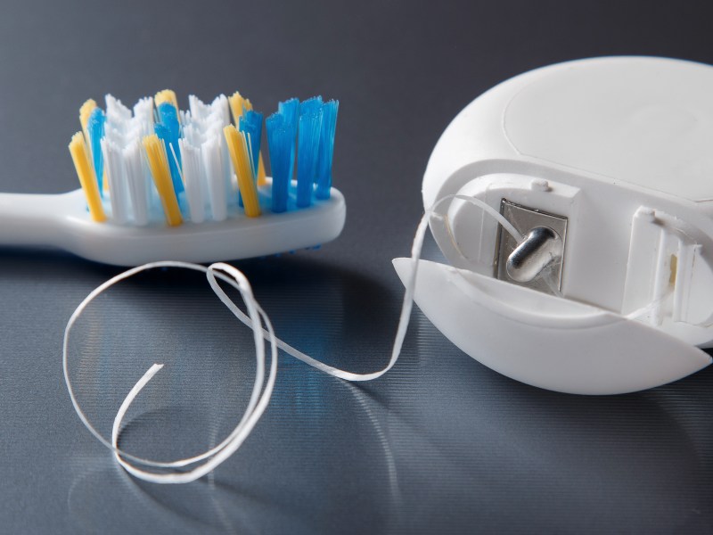 A toothbrush with blue and yellow bristles is next to an open container of dental floss on a gray surface.