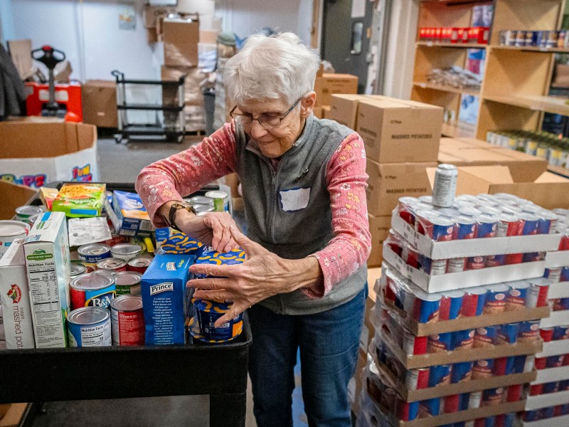 A woman organizes canned goods on a cart in a food pantry, surrounded by shelves and stacks of boxes.