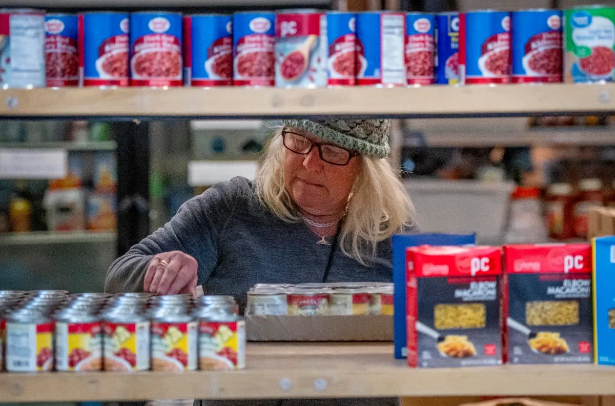 A person wearing glasses and a hat picks up a box in a store aisle stocked with canned goods and packaged food.