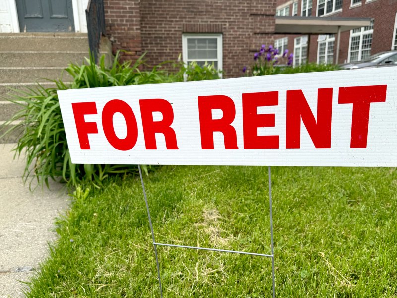 A red-and-white "for rent" sign on a grassy lawn in front of a brick building with stairs.