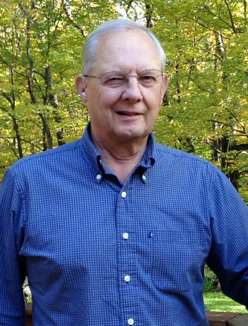 A man wearing glasses and a blue checkered shirt stands outdoors in front of trees with green and yellow leaves.