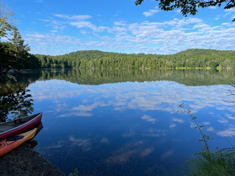 A calm lake reflects clouds and surrounding trees under a blue sky, with two kayaks partially visible at the shore in the lower left corner.
