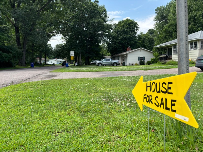 Yellow arrow-shaped sign reading "House for Sale" on grass points toward houses on a suburban street lined with trees and parked cars.