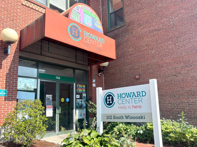 Entrance of Howard Center at 102 South Winooski, with a red awning, white sign, and surrounding plants in front of a brick building.