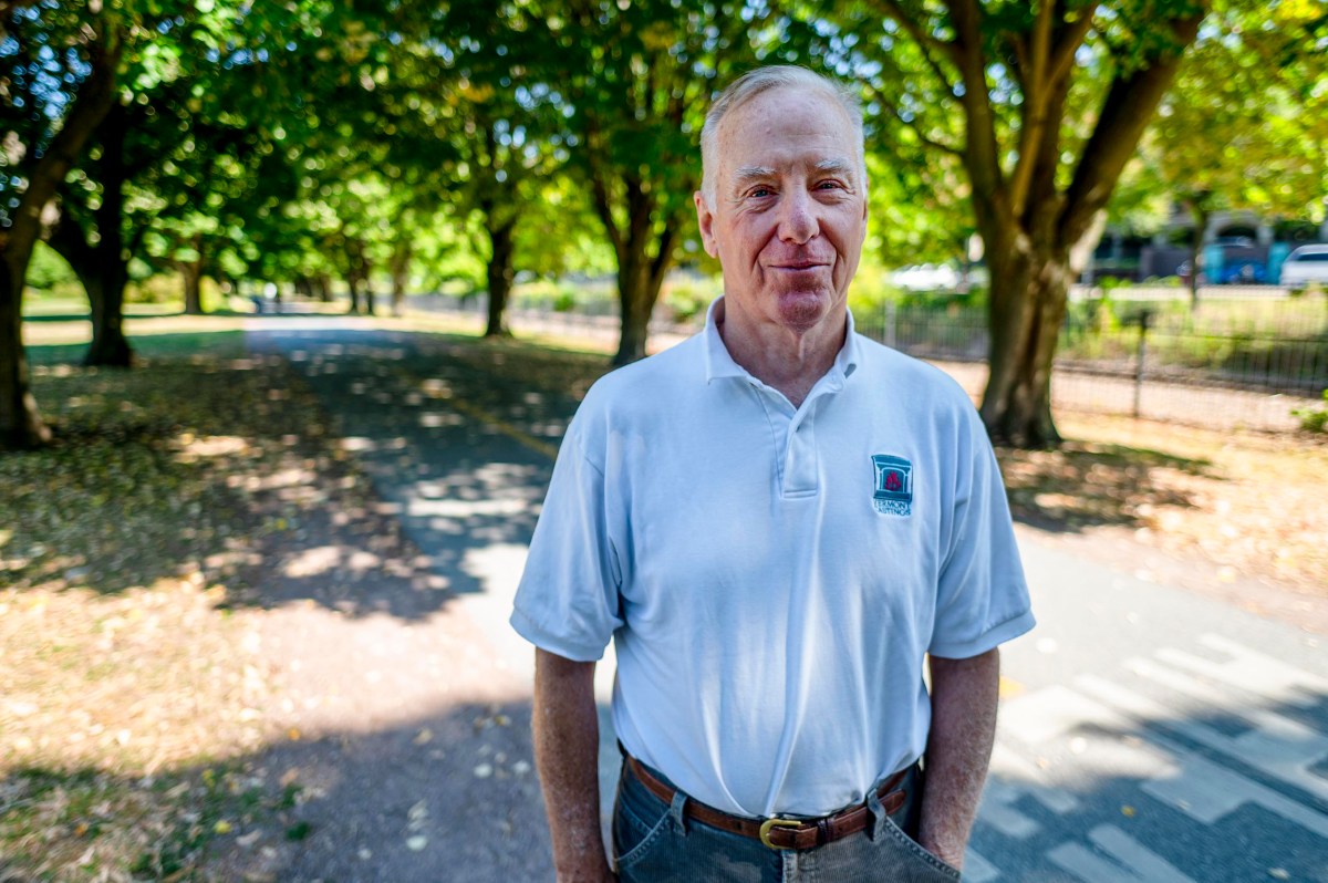An older man stands on a shaded park path, wearing a white polo shirt and gray pants, with trees and sunlight in the background.