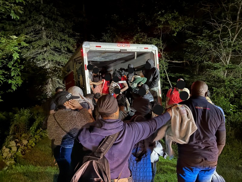 A group of people gather around the open back of a moving truck at night in a wooded area, with some loading or unloading items.