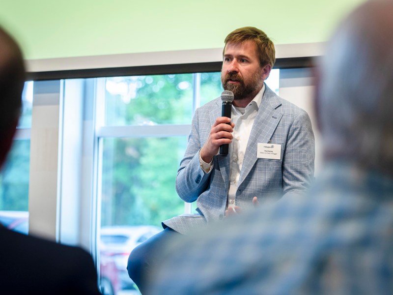 A man in a light blue suit jacket holds a microphone while speaking at an indoor event. Three people are seen from behind, listening to him.