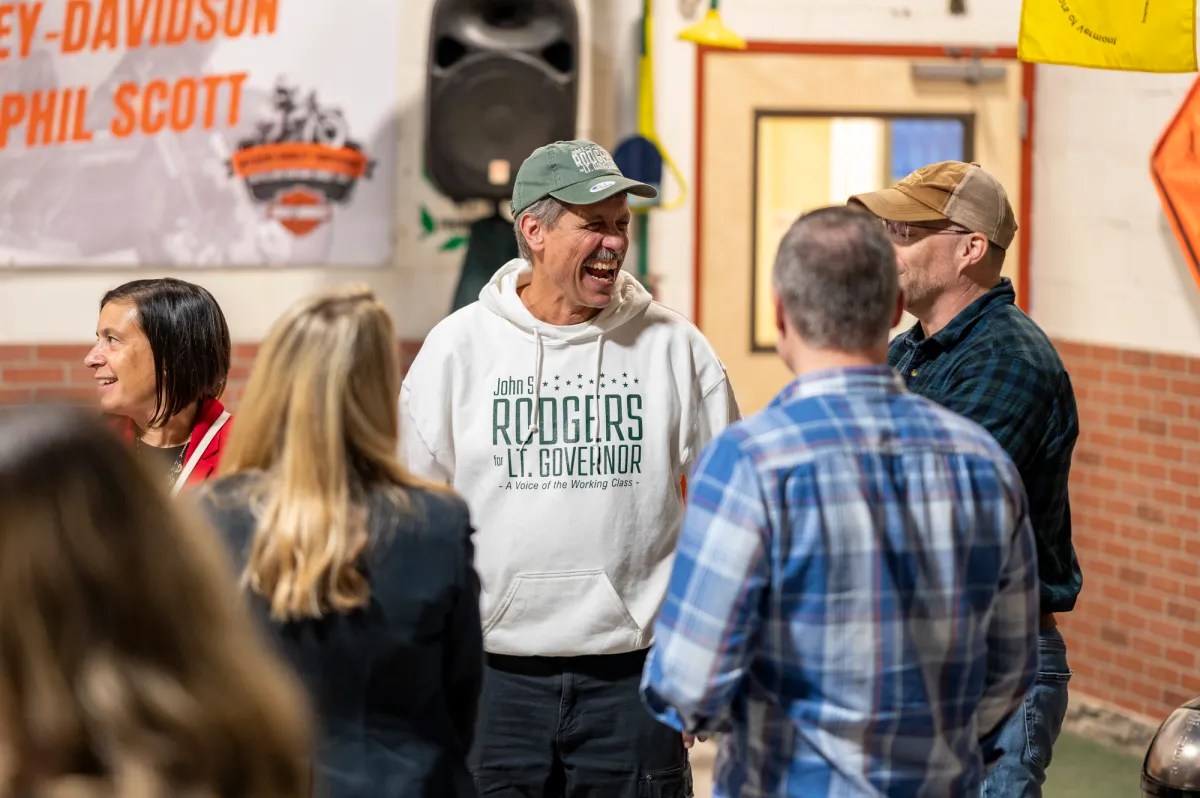 Group of people talking and smiling indoors, with one person wearing a hat and hoodie. Brick wall and banners are visible in the background.