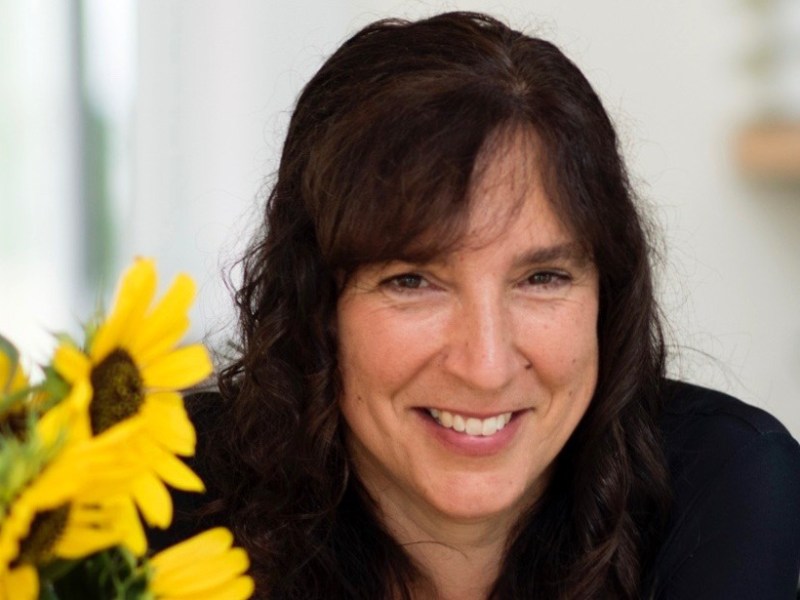 A woman with long, wavy dark hair smiles at the camera. Sunflowers are visible in the foreground on the left side.