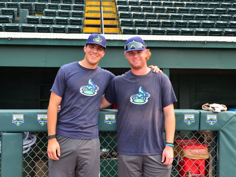 Two baseball players in matching team shirts and caps stand side by side on a field, smiling at the camera with empty stadium seats behind them.