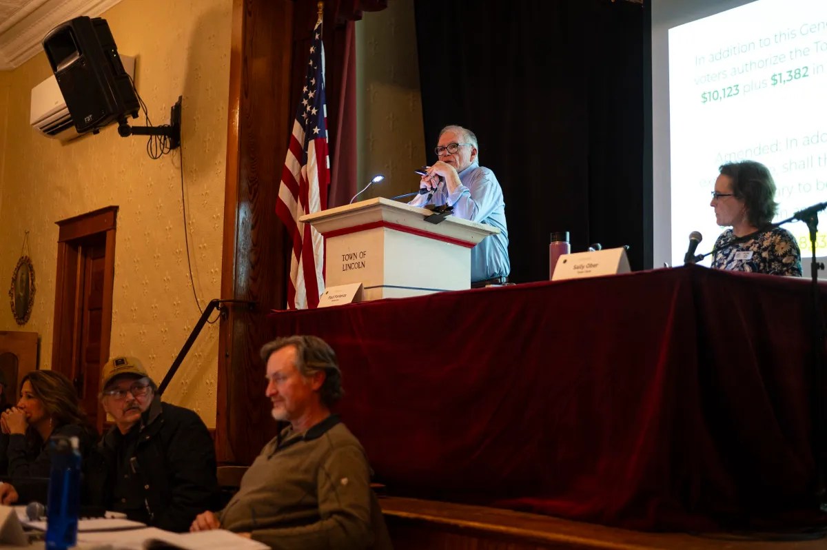 A person speaks at a podium labeled "Town of Lincoln" in an auditorium, with three people seated at a table nearby and an American flag in the background.