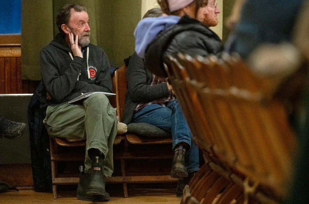A person sits in a wooden chair, hand on face, wearing a dark hoodie and green pants, in a room with others on similar chairs.